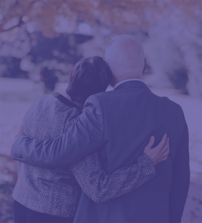 Elderly woman with black short hair has her head on the shoulder of elderly man looking away from the camera